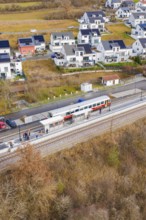 Train stops at a small train station next to modern houses surrounded by fields, historic event,