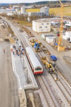 A train stops at a station under construction in an urban area, workers are present, historic