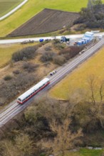 A train travels through a rural area along the railway tracks, surrounded by trees and fields,