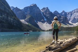 Female tourist standing on a log, admiring turquoise waters of moraine lake with canoers,