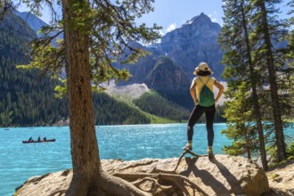 Female tourist standing on a rock by a tree, admiring the turquoise waters of moraine lake with