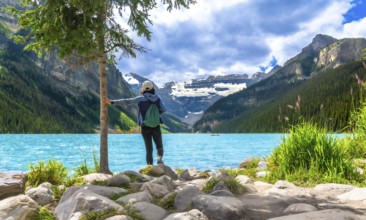 Female tourist standing by a tree on the rocky shore of turquoise lake louise, admiring the