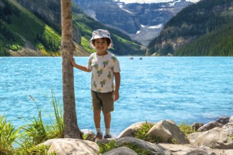 Young child standing by a tree on the rocky shore of turquoise lake louise, taking in the