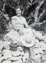 Portrait of young woman wearing summer dress sitting on stone wall holding straw hat, Malta,