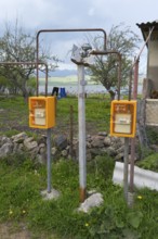 Two yellow gas meters stand on the side of a rural building with trees in the background, Sameba,