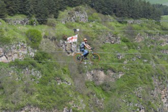 Person riding a bicycle on a rope in front of a wooded rocky landscape in the mountains, Dashbashi