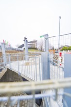 Modern outdoor area with steel railings and flags, building complex in the background, parking