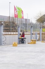 A worker sitting at a table next to a fence with assembled cables and boxes in an industrial area,