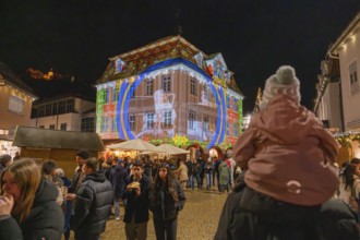 Families enjoy a night light projection onto a large building in a festive setting, Weihnachtsmark