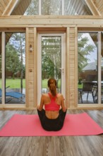 Back view of a woman sitting cross-legged on a pink yoga mat inside a wooden cabin. Outside, there