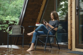 Barefoot woman sits on a chair at a table on a wet wooden porch and looks at a mobile phone. The