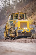 Yellow track construction machine runs on railroad tracks through wooded area, stuffing machine on