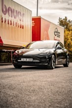 Black car in front of a truck sign on a sunny road in autumn, Deer E-Carsharing, Tesla Model 3,