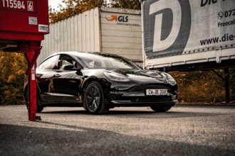 Black car between trucks in a parking lot with autumn scenery, Deer e-car sharing, Tesla Model 3,
