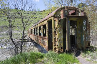 An abandoned, rusted drawbridge with graffiti in a green, hilly landscape on the riverbank,
