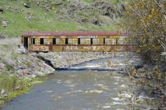 A rusted train car with graffiti stands over a flowing river in a green, hilly landscape, discarded