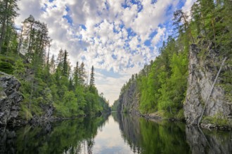 A calm lake lies in a wooded gorge, surrounded by tall rocks and reflecting the sky covered with