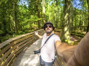 Photographer with hat and sunglasses taking a selfie while walking on a wooden path inside the