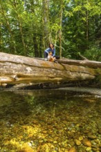 Mother and daughter sitting on a large fallen tree trunk over a clear creek in the tranquil