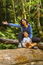 Cheerful mother and her son are sitting on a large fallen log in cathedral grove forest on
