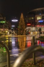 Large illuminated Christmas tree surrounded by colorful street lights in rainy city at night,