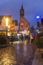 Christmas market with lights in front of a church, Christmas trees and people on wet cobblestones,