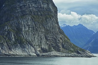 A massive rock juts out of the calm water, surrounded by a dramatic mountain backdrop under a