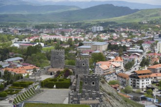 View of a historic castle with towers over a town with red roofs and mountain scenery, Rabati