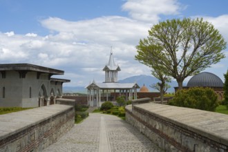 Architectural structure with tower and trees under a cloudy sky along a paved path, Rabati
