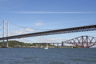 Two distinctive bridges over water with a small sailboat in the foreground, railway bridge and road