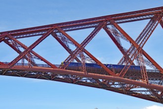 A train crosses a massive red steel bridge under a blue sky, Firth of Forth, Edinburgh, Scotland,