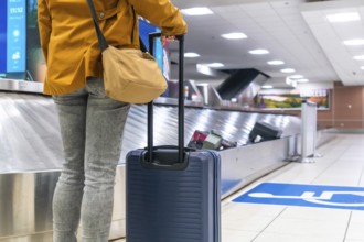 Tourist waiting at the baggage claim area in an airport, holding a suitcase while observing the