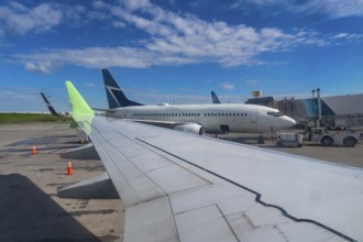 Airplane wing is overlooking airport runway with other aircraft parked and ground crew servicing