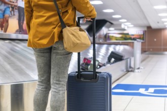Tourist is waiting for luggage at baggage claim carousel in airport terminal after arrival, holding