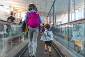 Mother and son are walking on a moving walkway at the airport, carrying backpacks and luggage as