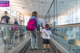 Mother and son with backpacks holding hands walking on a moving walkway in a modern airport