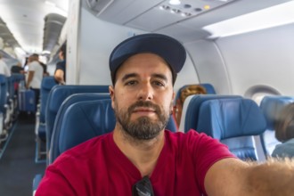 Bearded man capturing a selfie while seated in an airplane during a flight, with fellow passengers
