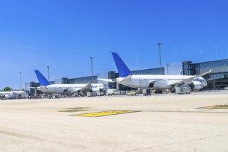 Two large passenger airplanes are parked at the gate of a modern airport on a sunny day, undergoing