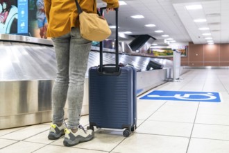 Traveler waiting by the baggage claim carousel at the airport, holding a trolley suitcase and a