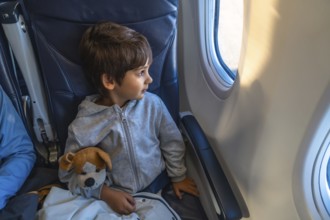 Curious child traveling by plane, clutching a plush toy while gazing out the window, marveling at
