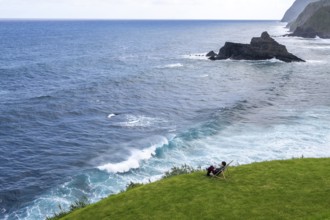 Woman sitting comfortably in deckchair in a meadow and watching waves in the sea, Madeira, Portugal