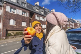 Two young women laughing and sharing a healthy snack on a sunny urban street, bundled in coats and