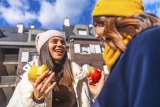 Two cheerful women friends enjoying a healthy apple snack and talking outdoors on a sunny winter