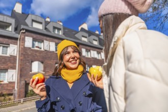 Two young women laugh and chat on a sunny autumn city street, sharing fresh apples and cozying up