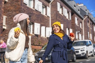 Smiling young women friends walking and talking on a city street, enjoying a healthy snack of