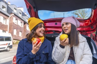 Two cheerful women sit in a car trunk on a winter road trip, sharing apples, laughing and talking