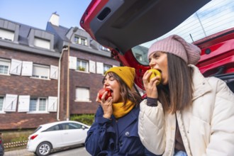 Two young women sit in an open car trunk on a city street, laughing and sharing fresh apples during