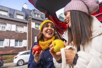 Two cheerful young women laughing while wearing hats and holding apples, standing by an open car