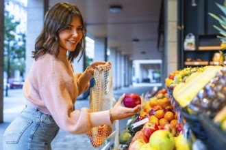 Young woman smiling while adding fresh red apples to her reusable mesh bag at an outdoor or street