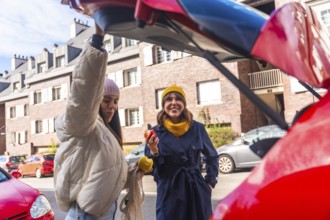 Two cheerful young women are holding apples and smiling, standing together by the open trunk of a